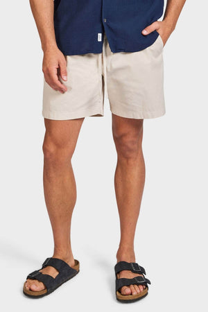 Dressed in a navy shirt and Academy Brand Oahu Short in light beige cotton twill with black buckle sandals, a person stands with one hand in their pocket against a plain white background, radiating easy vintage style.