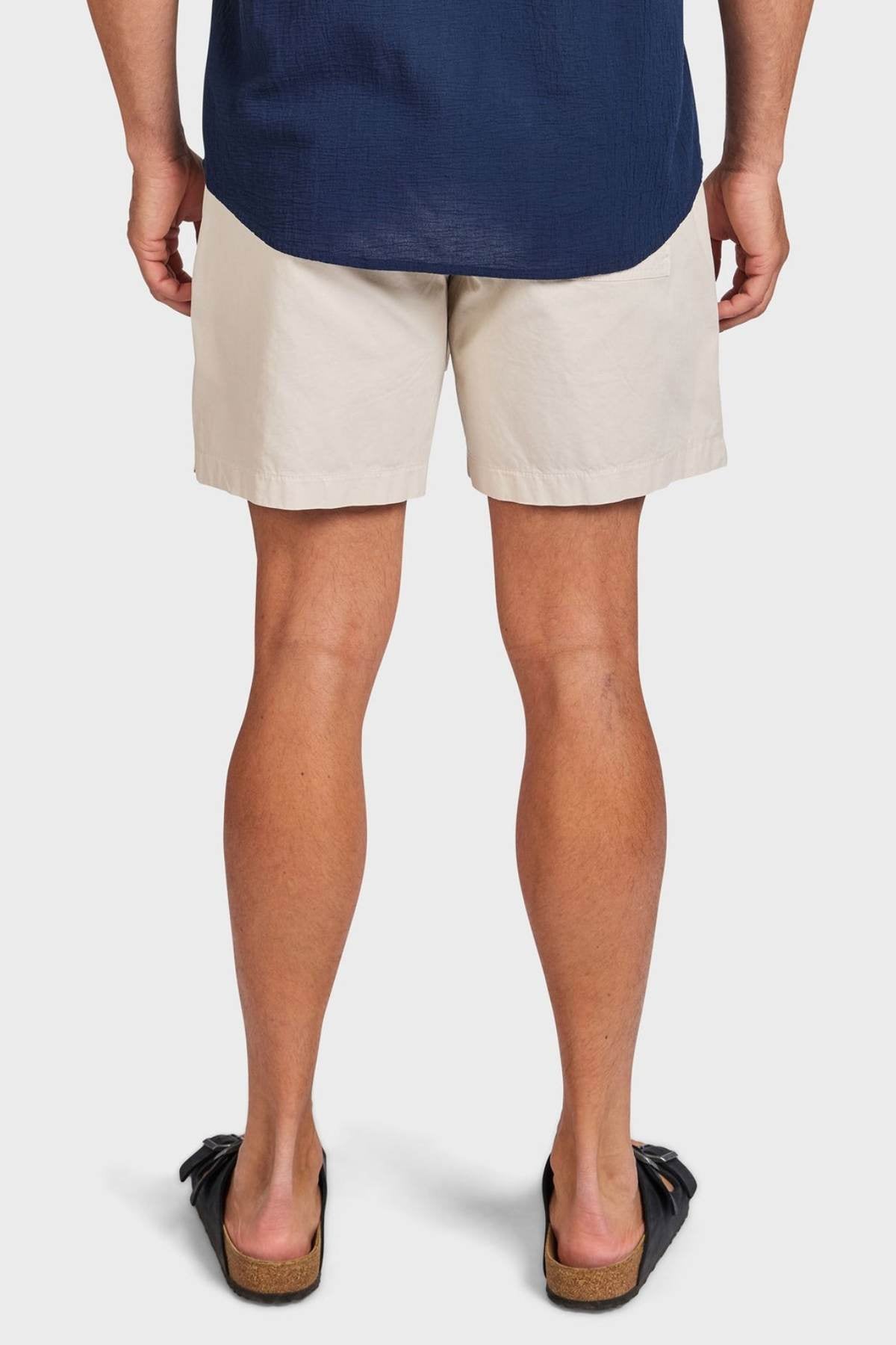 A person stands facing away, wearing a navy blue shirt, Academy Brand Oahu Short in vintage cotton twill, and black slide sandals with cork soles, set against a plain white background.