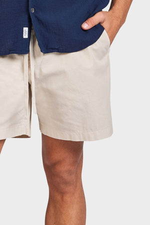 A person stands with one hand in the pocket of Academy Brand Oahu Short in light beige cotton twill, wearing a navy blue shirt, with part of the arm and upper legs visible against a plain white background.