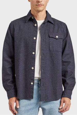 Clad in an Academy Brand Oakland Overshirt in dark grey, made from premium yarn-dyed twill and 100% cotton, a person showcases its design which includes a chest pocket on the left side with a button closure. The stylish look is complemented with light blue jeans and a white t-shirt underneath, making the ensemble pop against the plain white background.