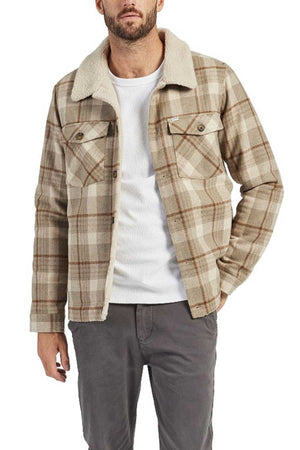 A man wearing the Academy Brand Quincy Jacket, a tan and cream plaid wool blend with faux shearling lining, over a white shirt and gray pants, stands against a white background.