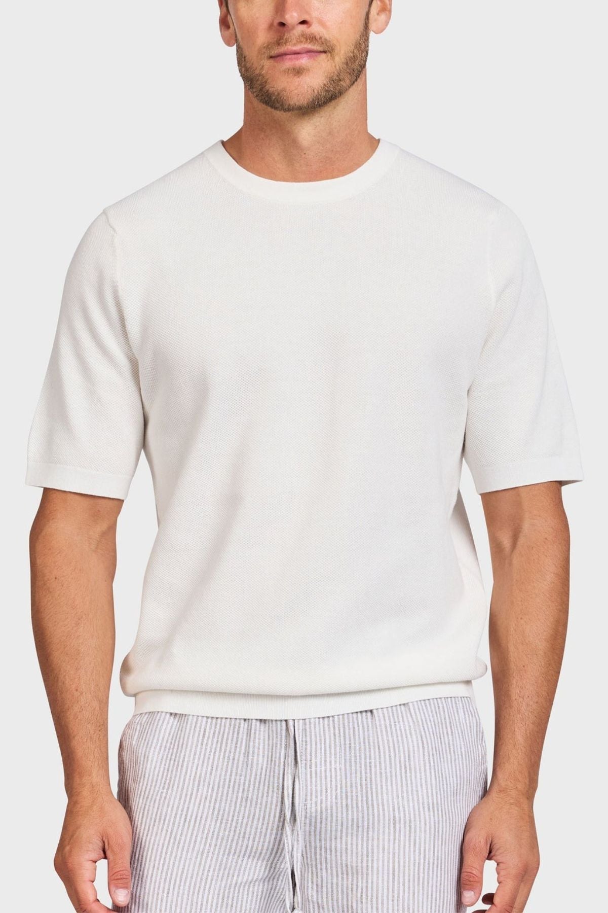 A man in a plain white Academy Brand Riley Knit Tee, crafted from lightweight, breathable fabric, paired with light gray striped pants stands against a light background. The minimalist and casual outfit highlights the premium textured stitch of his attire.
