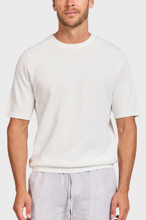 A man in a plain white Academy Brand Riley Knit Tee, crafted from lightweight, breathable fabric, paired with light gray striped pants stands against a light background. The minimalist and casual outfit highlights the premium textured stitch of his attire.