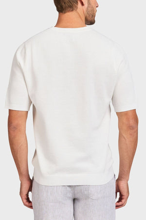 A man is seen from behind in an Academy Brand Riley Knit Tee, a white short-sleeve sweater made of lightweight breathable fabric, paired with light gray striped pants against a plain white background.