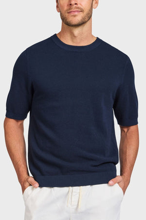 A man in a dark blue Academy Brand Riley Knit Tee and light pants stands with hands in pockets against a white background, highlighting the premium textured stitch that adds elegance and style.