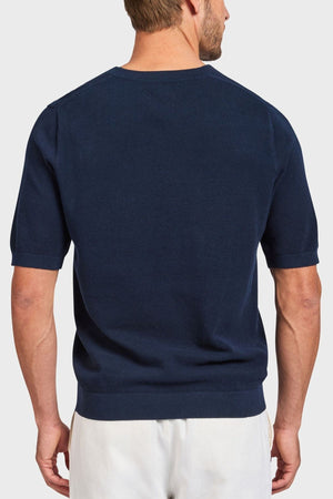 A man faces away from the camera, highlighting an Academy Brand Riley Knit Tee in navy blue made from breathable fabric, paired with crisp white pants against a plain light gray background.