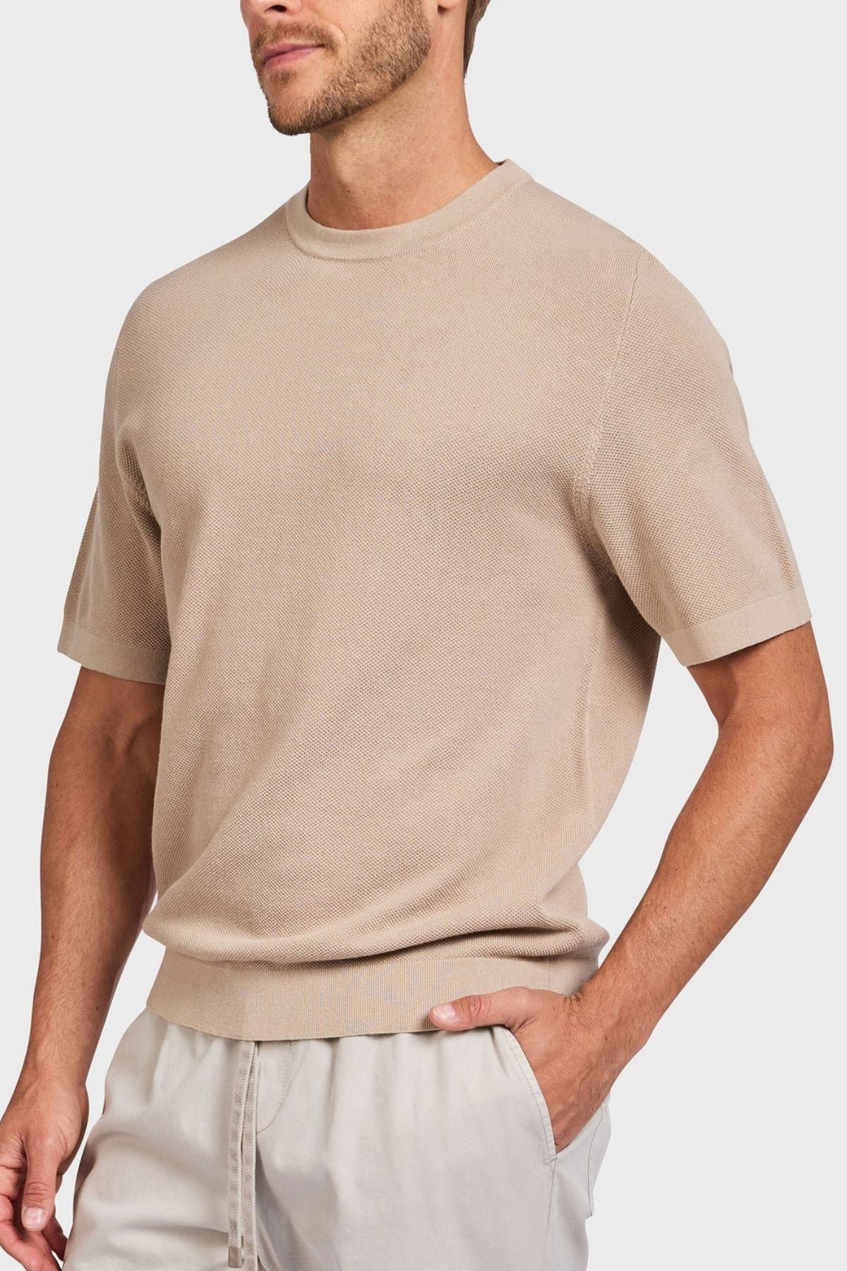 A person in a light beige Academy Brand Riley Knit Tee, made from lightweight breathable fabric, pairs it with light gray pants against a plain background, showcasing comfortable casual style with premium textured stitch detail.