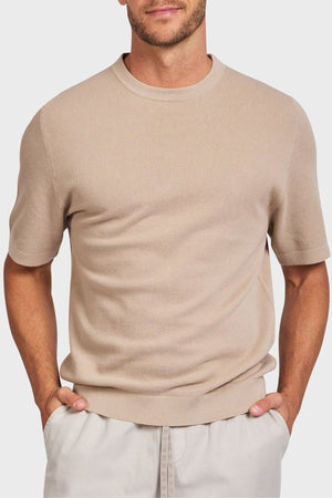 A person stands against a plain background, hands in pockets, wearing the Academy Brand Riley Knit Tee, a beige short-sleeve sweater made from lightweight breathable fabric, paired with light gray pants.