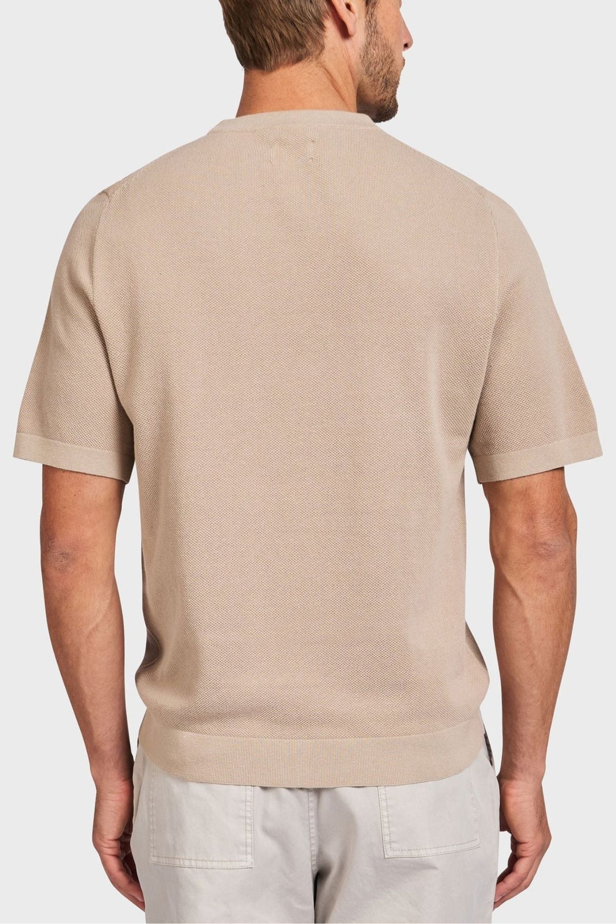 A person clad in an Academy Brand Riley Knit Tee, a beige short-sleeve polo shirt made of lightweight breathable knit fabric, and light gray pants is shown from the back against a plain background.