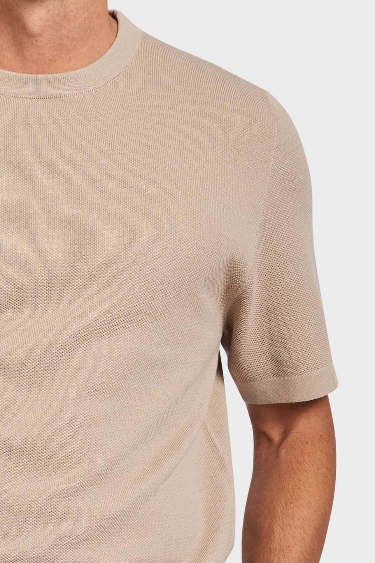 A person wearing an Academy Brand Riley Knit Tee is pictured. The image highlights the premium textured stitch and fit of the beige short-sleeve crew neck sweater against a plain white background, emphasizing its lightweight and breathable fabric.