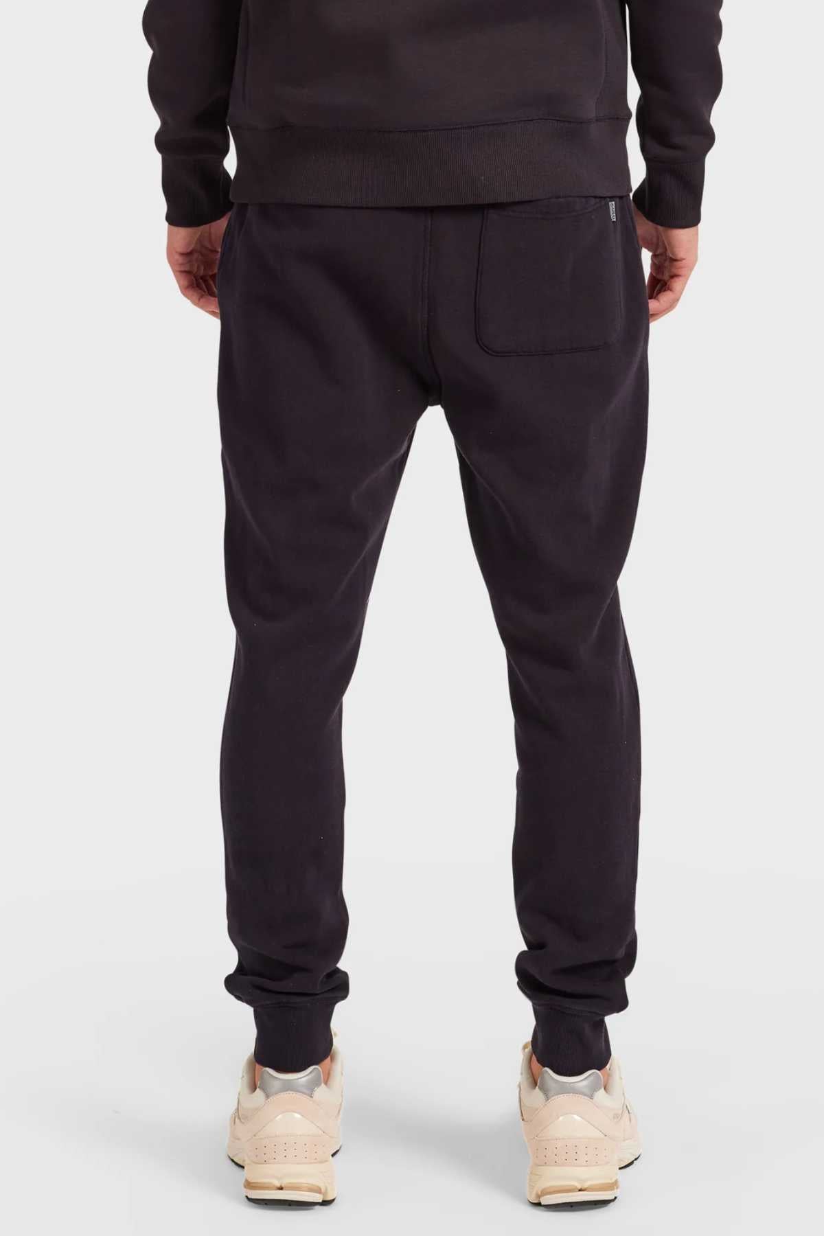 A person is seen from the back wearing Academy Brand Sweat Pant tracksuits in black with elastic waistbands, styled with light beige sneakers. Made from premium brushed fleece, these pants display a visible back pocket, set against a plain white backdrop.