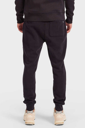 A person is seen from the back wearing Academy Brand Sweat Pant tracksuits in black with elastic waistbands, styled with light beige sneakers. Made from premium brushed fleece, these pants display a visible back pocket, set against a plain white backdrop.