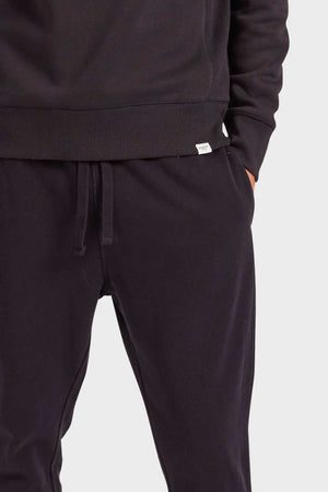 Wearing a black hoodie and Academy Brand Sweat Pant featuring an elastic waist and premium brushed fleece, a person slips their hands into the pockets. Only their torso and upper legs are visible against a plain white background.