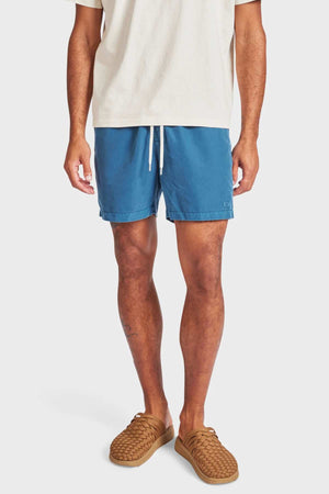 A person wearing a cream T-shirt, Academy Brand Teamster Boardy blue recycled polyester swim shorts, and tan woven slip-on shoes stands against a plain light background with their face not visible.