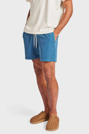 A person stands with hands in pockets, wearing a white T-shirt, Academy Brand Teamster Boardy blue quick dry swim shorts, and tan woven slip-on shoes against a plain light background.