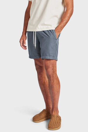 A man wears a cream short-sleeve shirt, Academy Brand Teamster Boardy swim shorts in blue-gray, and tan woven slip-on shoes, posing with one hand in his pocket against a plain white background.