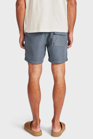 A person stands facing away, wearing the Academy Brand Teamster Boardy tailored fit blue-gray quick dry swim shorts with a back pocket, beige t-shirt, and tan sandals against a plain white background.