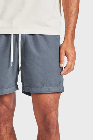 A person wears the Academy Brand Teamster Boardy—a tailored fit blue drawstring short made from recycled polyester—paired with a beige t-shirt, shown waist to mid-thigh with one arm relaxed at their side.