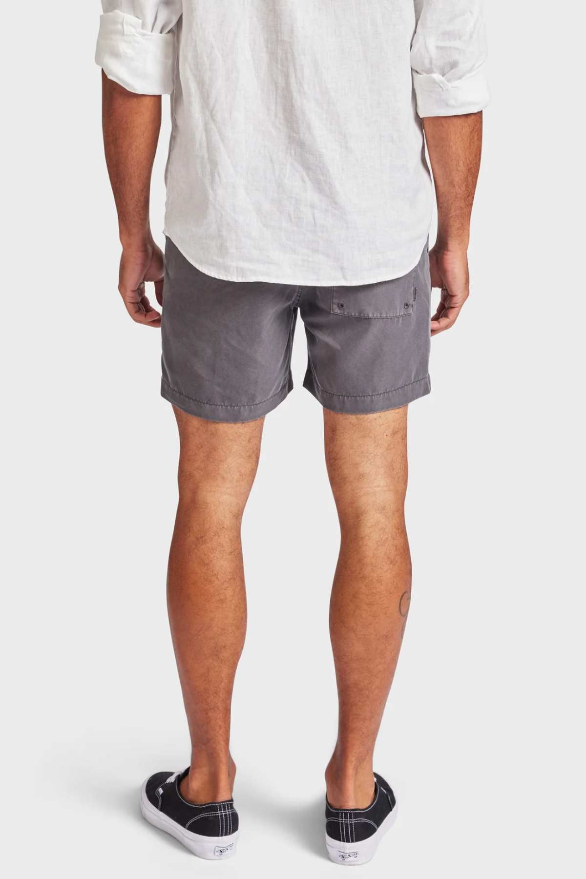 The person is wearing the Academy Brand Teamster Boardy, a white long-sleeve shirt, and tailored-fit gray shorts made from recycled polyester. They're standing against a plain background, paired with black slip-on shoes that feature white soles.