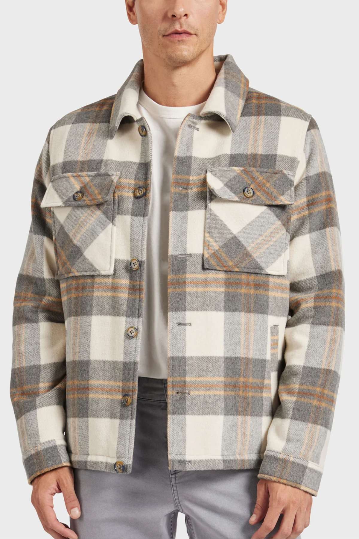 A person wears the Academy Brand Tulsa Jacket in plaid with beige, gray, and tan over a white shirt. This wool blend jacket has button-up front, two chest pockets, and quilted lining for warmth. The look is completed with gray pants on a plain white background.