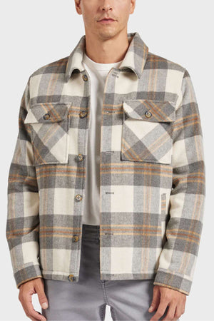 A person wears the Academy Brand Tulsa Jacket in plaid with beige, gray, and tan over a white shirt. This wool blend jacket has button-up front, two chest pockets, and quilted lining for warmth. The look is completed with gray pants on a plain white background.