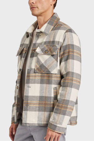Someone is wearing an Academy Brand Tulsa Jacket, a wool blend in plaid with brown, gray, and cream hues. It has a quilted lining, two chest pockets, and a collar. The person is posed against a plain gray backdrop, visible only from the upper body.