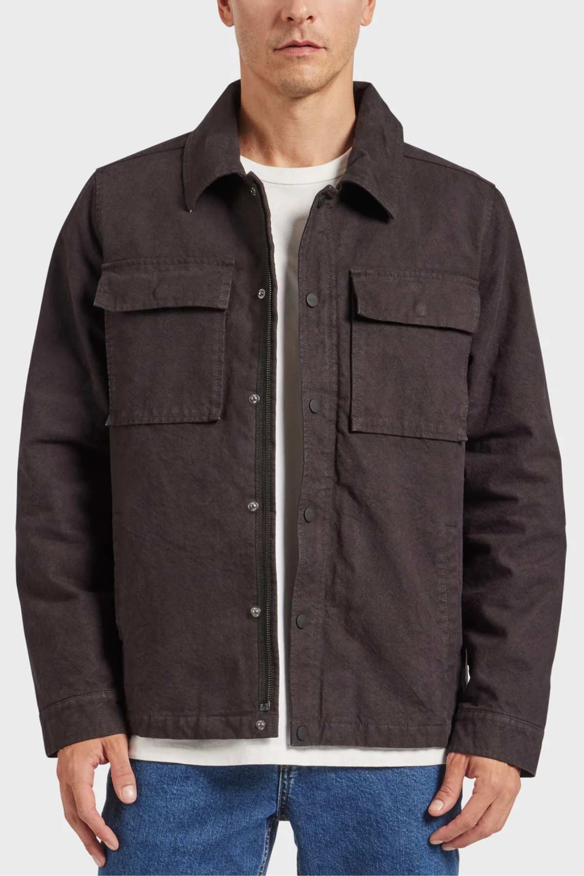 A person is wearing the Academy Brand Tulsa Jacket, a dark button-up made of cotton canvas featuring a collar and two front chest pockets, over a white shirt. They have paired it with blue jeans. The background is plain and light-colored.
