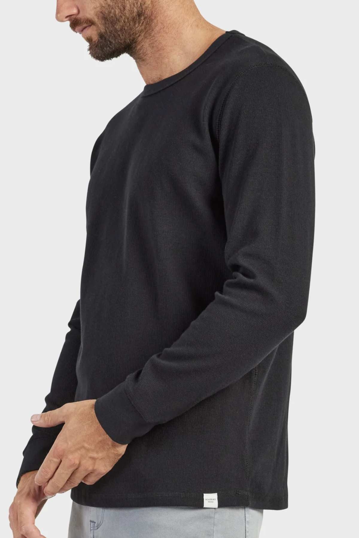 An individual is dressed in a black, long-sleeve Academy Brand Workers Crew shirt, featuring French rib construction. They are posed sideways with their head slightly angled downward, showcasing short hair and a beard. Their hands rest comfortably in front of them against a simple white backdrop.