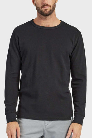 A man with a beard dons an Academy Brand Workers Crew long-sleeve, black crew neck shirt made from French rib fabric, paired with light gray pants. He stands against a plain white background.