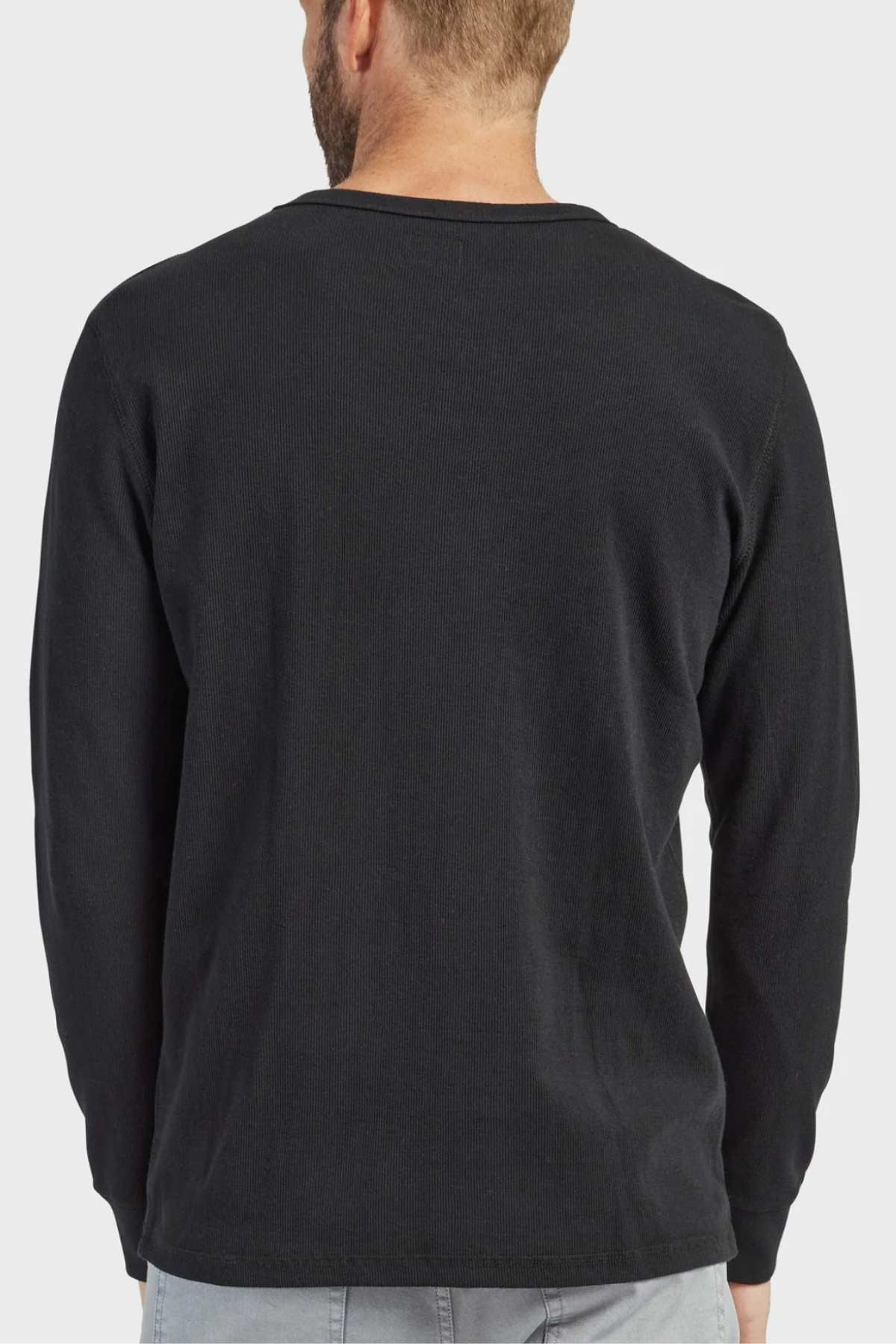 A man with light hair is standing with his back to the camera, dressed in a long-sleeve crew neck black shirt and light grey pants. The background is plain white, showcasing the French rib construction of his Academy Brand Workers Crew apparel.