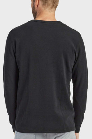 A man with light hair is standing with his back to the camera, dressed in a long-sleeve crew neck black shirt and light grey pants. The background is plain white, showcasing the French rib construction of his Academy Brand Workers Crew apparel.