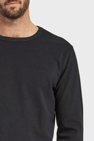 A close-up of a man wearing an Academy Brand Workers Crew, a dark long-sleeve top featuring a round neckline. The image captures him from the shoulders to the lower face, emphasizing the French rib construction and the simplicity of his attire against a plain white background.