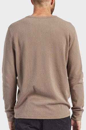 A person with short hair wears the Academy Brand Workers Crew, a durable beige long-sleeve crew neck sweater, and dark pants, standing with their back to the camera against a plain, light gray background.