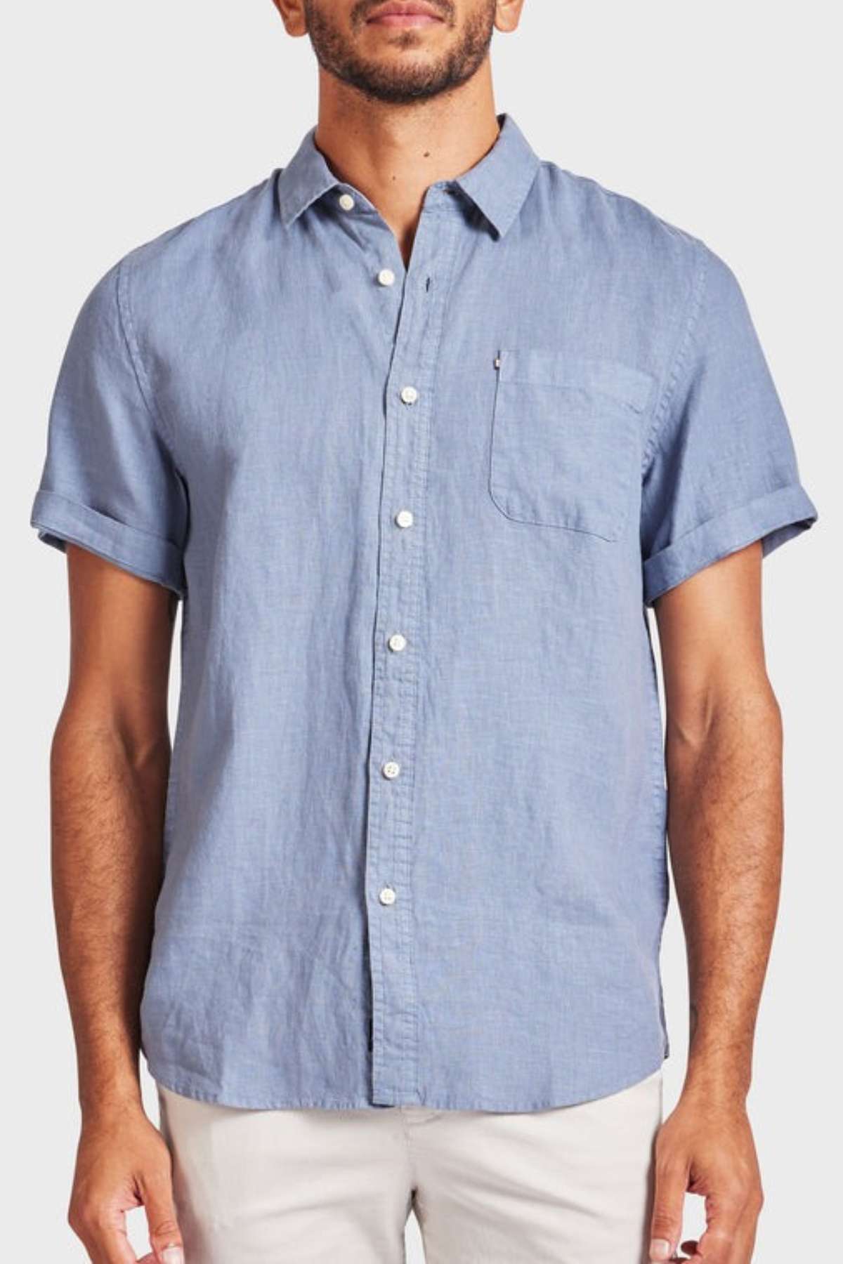 A man wears the Academy Brand Hampton Linen Short Sleeve Shirt in light blue, featuring a chest pocket and white buttons, paired with light-colored pants against a plain background.