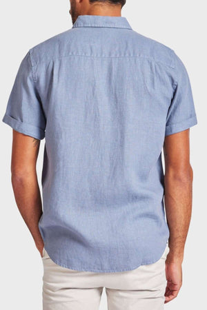 A man is seen from the back wearing a light blue Academy Brand Hampton Linen Short Sleeve Shirt and white pants, with his hands in his pockets against a plain light background.
