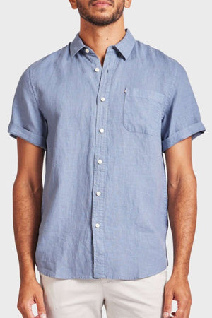 A man wears the Academy Brand Hampton Linen Short Sleeve Shirt in light blue, featuring a chest pocket and white buttons, paired with light-colored pants against a plain background.