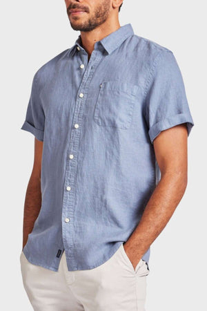 A man wears the Academy Brand Hampton Linen Short Sleeve Shirt in light blue, featuring a chest pocket, paired with beige pants, standing with hands in pockets against a plain background.