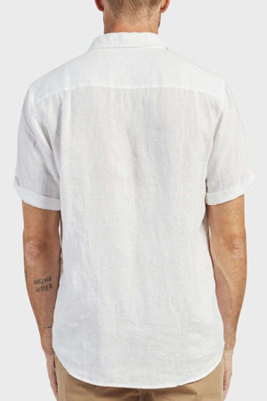 A man with a tattoo on his left forearm is seen from behind wearing the Academy Brand Hampton Linen Short Sleeve Shirt in white with tan pants, standing against a plain light background.