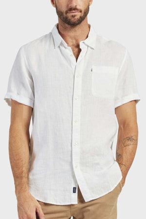 A man wears the Academy Brand Hampton Linen Short Sleeve Shirt—white, button-up with a chest pocket—and beige pants, standing against a light gray background. Tattoos are visible on his left arm.