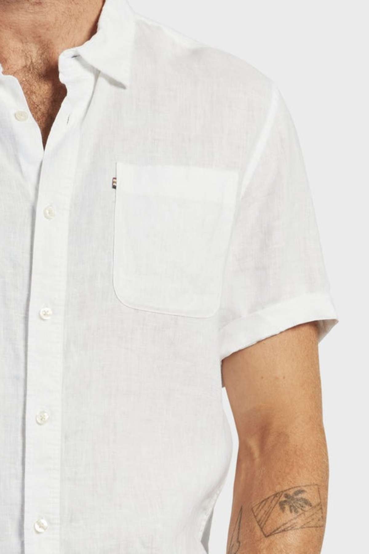 A person wears the Academy Brand Hampton Linen Short Sleeve Shirt featuring a chest pocket. The photo captures the upper body, displaying part of the left chest, arm, and a geometric forearm tattoo. Made from premium linen for comfort and style.