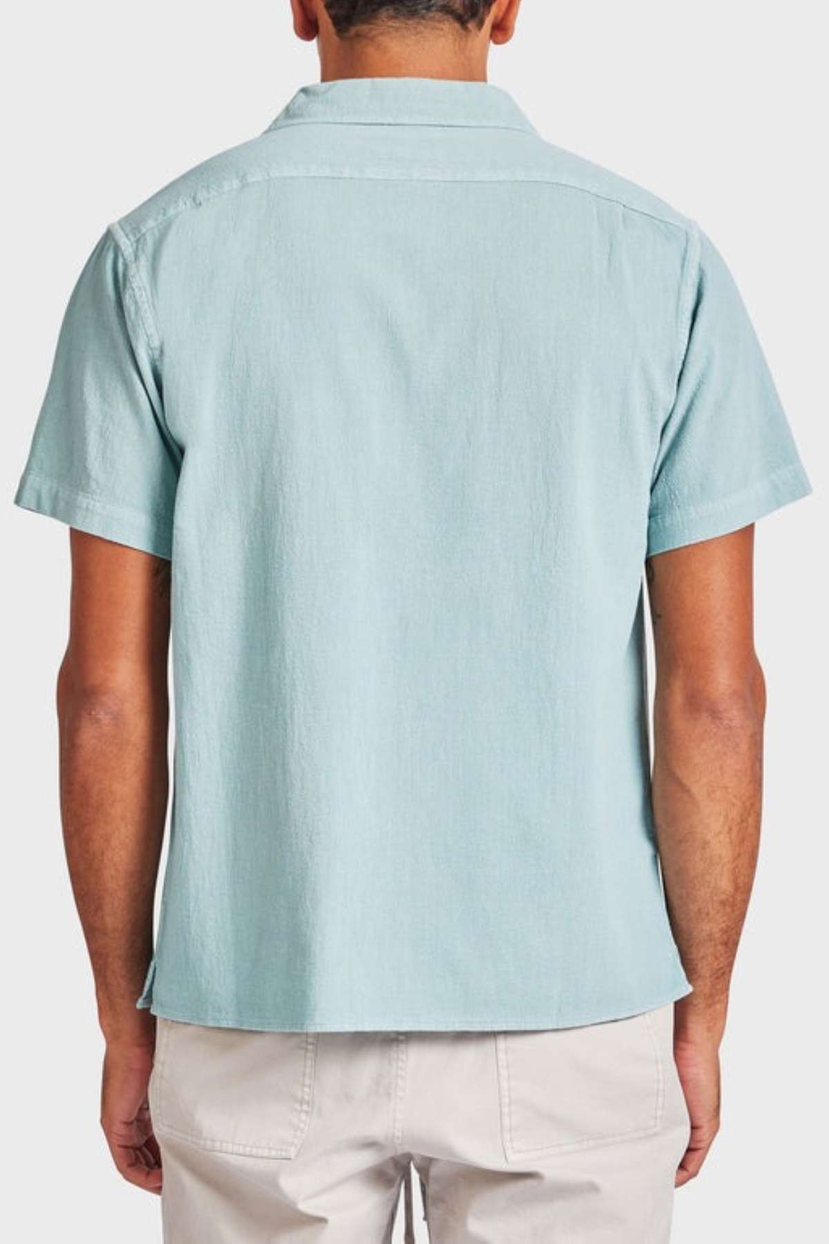 Rear view of a person wearing the Academy Brand Jed Short Sleeve Shirt in light blue textured cotton with a classic revere collar, paired with light beige pants. The individual stands gracefully against a plain white background.