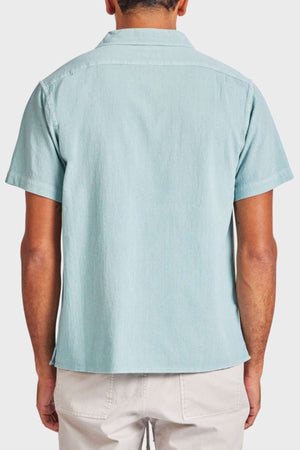 Rear view of a person wearing the Academy Brand Jed Short Sleeve Shirt in light blue textured cotton with a classic revere collar, paired with light beige pants. The individual stands gracefully against a plain white background.