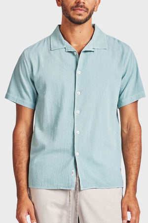 A man stands against a plain background wearing the Academy Brand Jed Short Sleeve Shirt, which features a revere collar and a relaxed fit in light blue. He pairs it with light-colored pants for a casual look.