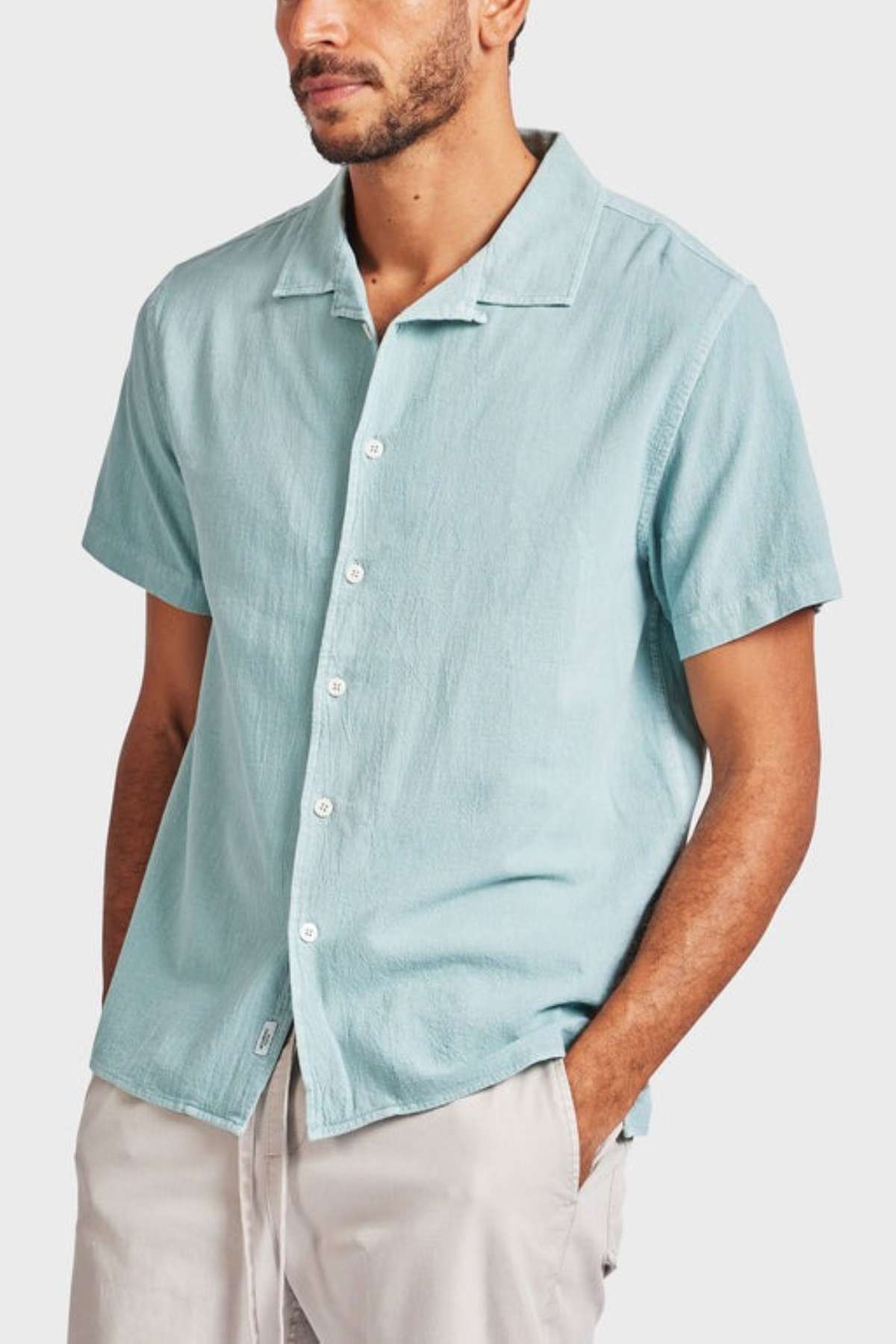 A person wearing the Academy Brand Jed Short Sleeve Shirt in light blue with a revere collar and light-colored pants stands against a plain background. The shirt's textured fabric adds subtle depth as their left hand rests casually in their pocket.