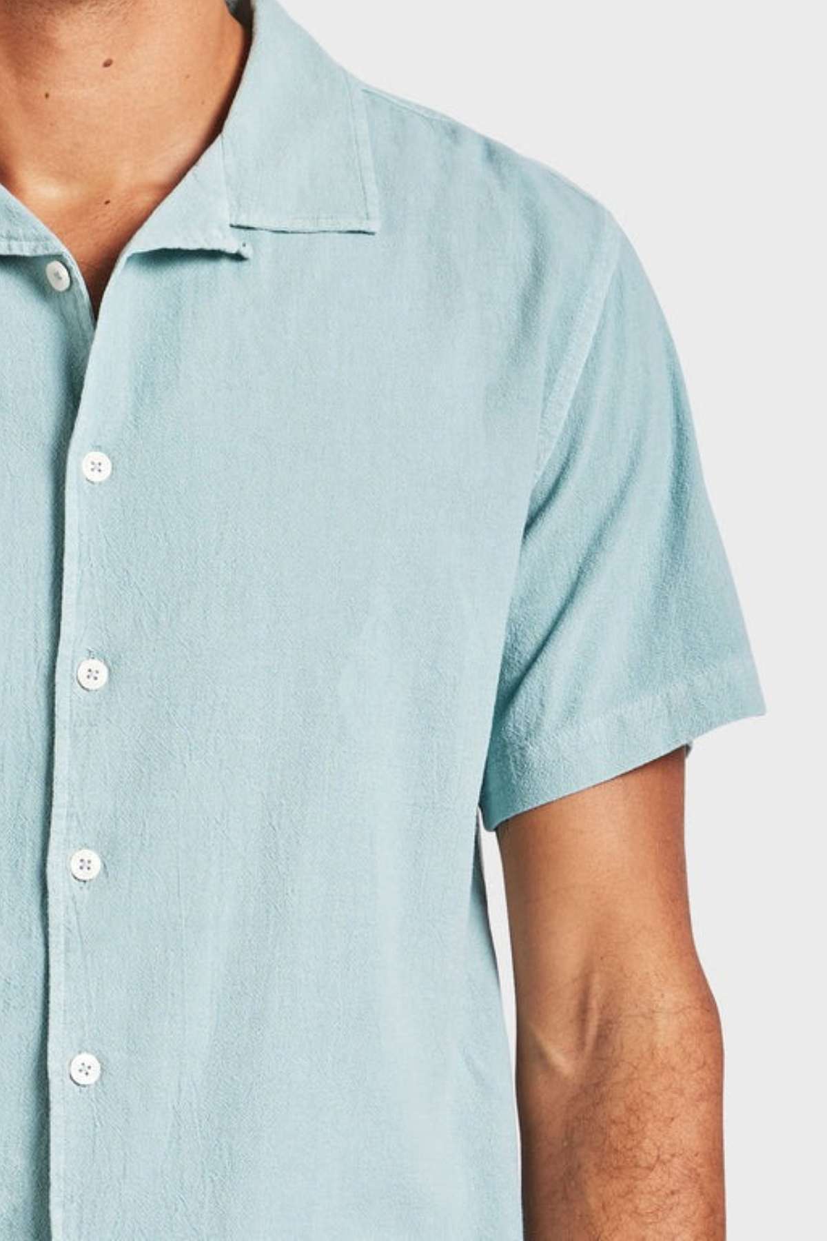Someone wears the Academy Brand Jed Short Sleeve Shirt in light blue, featuring soft cotton, a textured fabric, short sleeves, and a revere collar. The shoulders are cropped in the image set against a plain white background.
