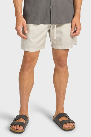 A person wears light beige Academy Brand Oahu Short cotton twill shorts with a relaxed volley silhouette, paired with a gray textured shirt and black slide sandals. The image is cropped to show only the lower torso and legs.