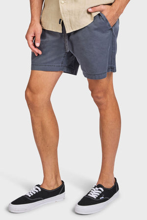 Wearing a beige shirt, Academy Brand Oahu Short, and black-and-white sneakers, a person stands with one hand in their pocket against a plain white background, exuding vintage charm.
