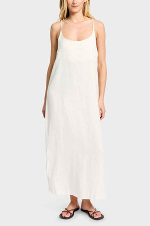 A person stands against a plain background wearing the Academy Women Essential Linen Slip Dress, a long white linen garment with adjustable straps and subtle side splits. The outfit is completed with black flip-flops.