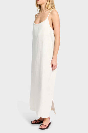 A person wearing the Academy Women Essential Linen Slip Dress in cream stands against a plain background. This simple, sleeveless dress includes adjustable straps and a subtle texture, paired with black sandals for an effortlessly chic look.