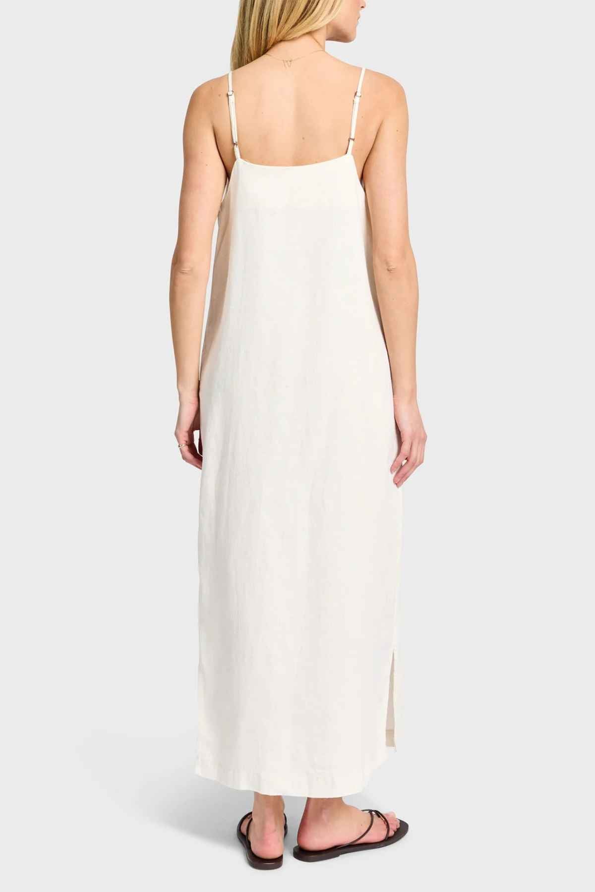 A person is wearing the Academy Women Essential Linen Slip Dress, a long, cream-colored piece with adjustable straps that gracefully drapes over their figure and features subtle side splits. They are facing away from the camera on a light gray background, paired with black sandals.